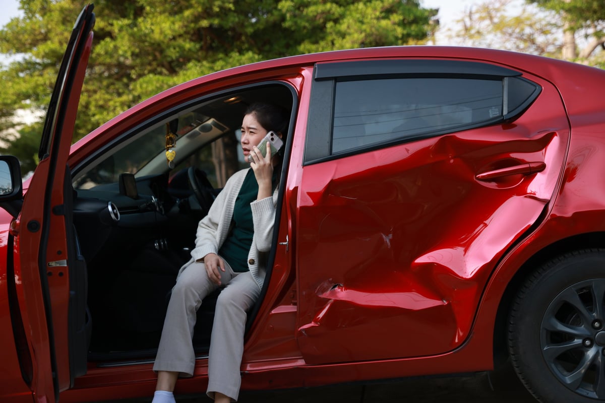 A female motorist standing on the side of the road after a car accident, assessing the damage to her vehicle. She confidently calls for roadside assistance.