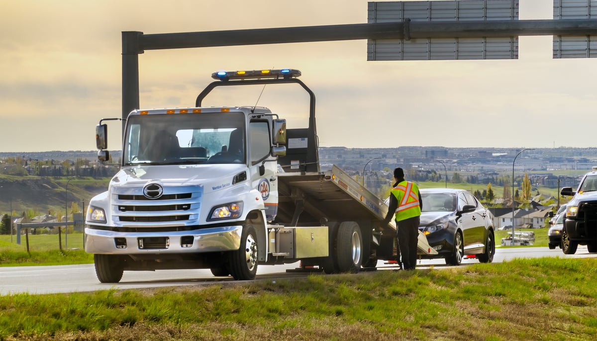 A tow truck driver assists a broken-down sedan on a highway shoulder during a clear day