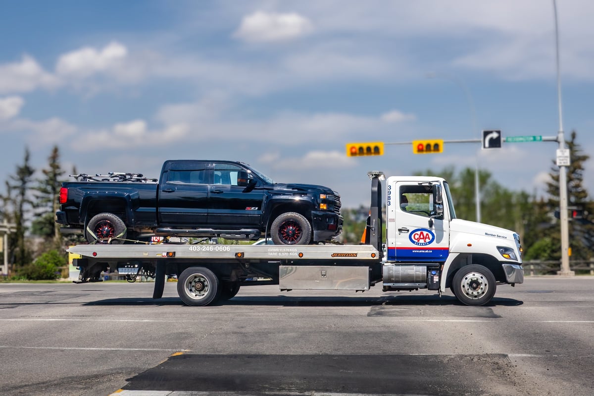 White CAA flatbed tow truck transporting a black pickup truck through a city intersection