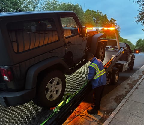 Tow truck operator loading a dark SUV onto a flatbed tow truck on a residential street