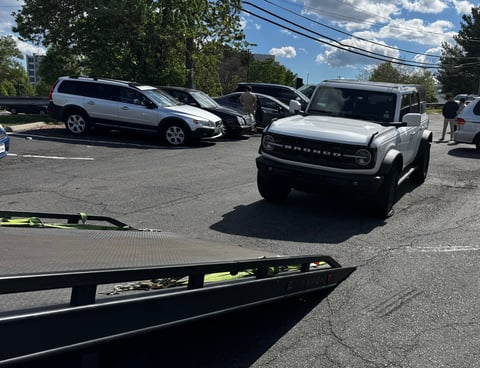 White SUV parked in a lot with other vehicles, power lines and trees visible under a cloudy sky