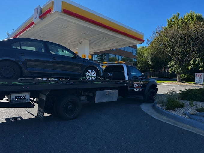 Tow truck carrying a dark sedan at a gas station with red and yellow canopy on a sunny day