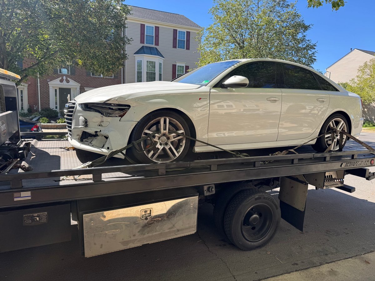 White damaged car loaded on a black tow truck with residential houses in the background