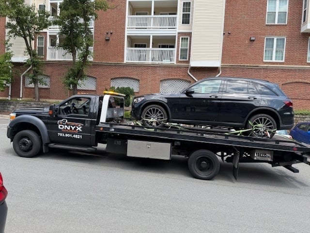 Black tow truck with ONYX branding transporting a dark sedan on flatbed in front of brick apartment building