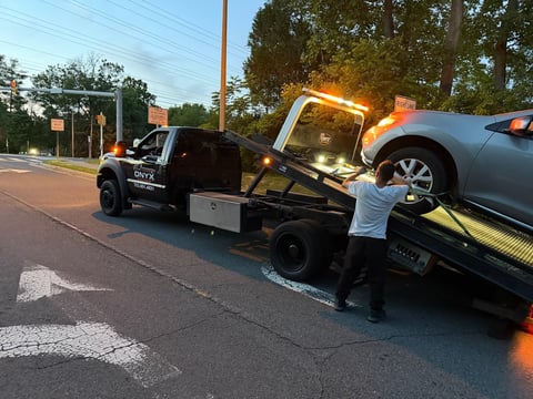 Black tow truck loading a silver sedan onto its flatbed during daytime on a residential street