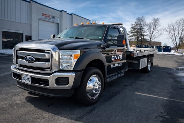 Black Ford F-450 Onyx flatbed truck with amber warning lights parked in a lot near an industrial building.