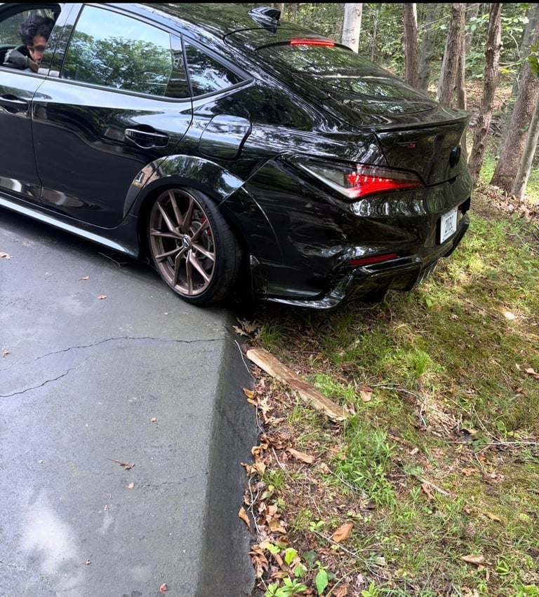 Black sport sedan parked on driveway next to wooded area, rear bumper visible with red taillights