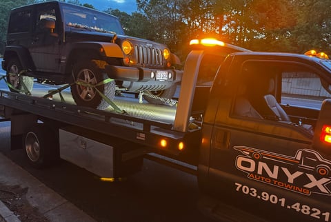 Black tow truck with orange/yellow headlights carrying a dark Jeep on its bed, during dusk or early evening
