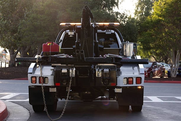 Black utility truck with hydraulic lift arm and equipment mounted on back, parked on street with trees in background