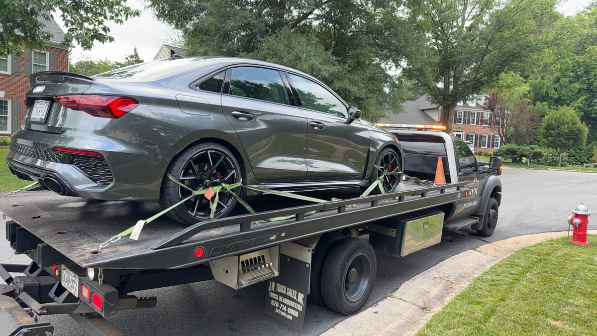 Gray sedan being towed on a flatbed truck on a residential street lined with trees and brick houses