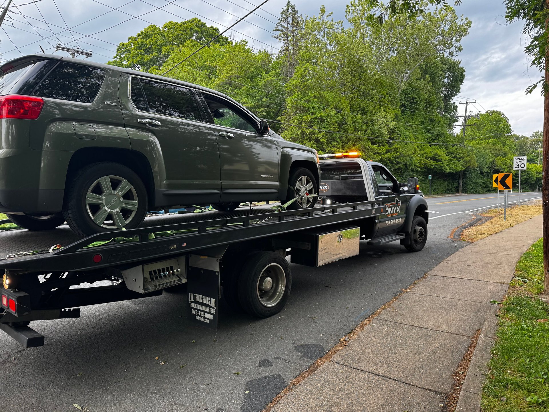 A black tow truck carrying a dark green SUV on a residential street lined with trees and power lines