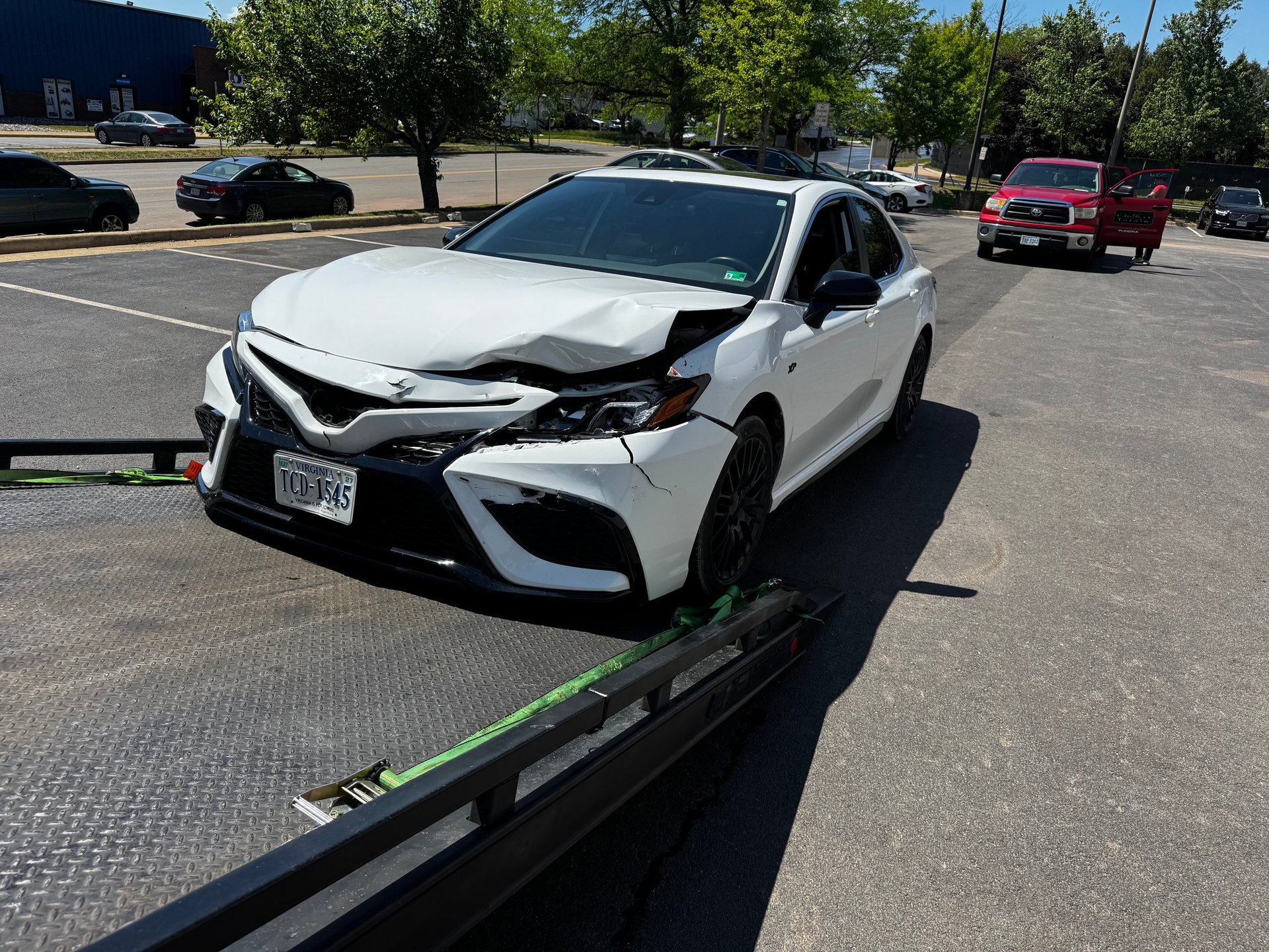 White Toyota Camry with front-end damage being towed on a flatbed truck in a parking lot