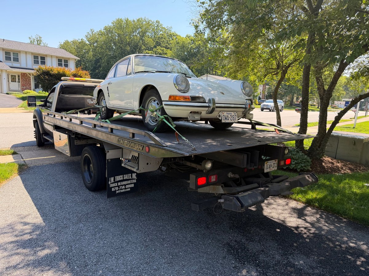 White vintage Porsche being towed on a flatbed truck in a residential neighborhood with trees