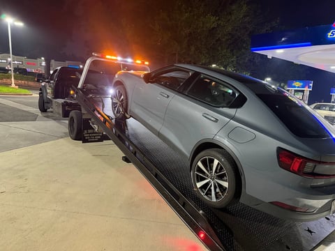 Silver sedan being loaded onto a tow truck at night in a parking lot with gas station in background