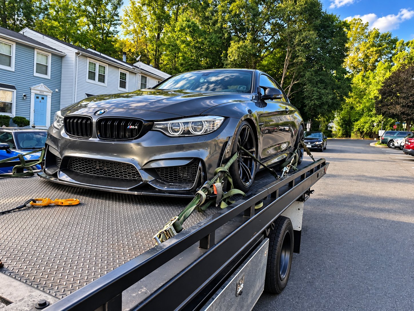 Dark gray BMW M4 sports car being transported on a flatbed tow truck in a residential neighborhood