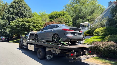 Gray sedan being towed on a flatbed truck in a residential street with green trees