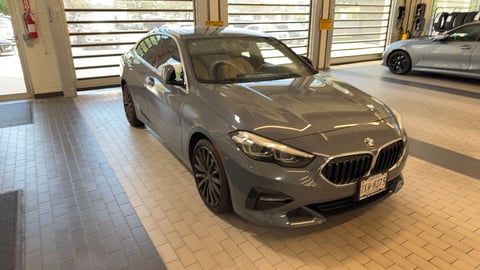 Silver BMW sedan displayed in a modern showroom with glass doors and another vehicle visible in the background