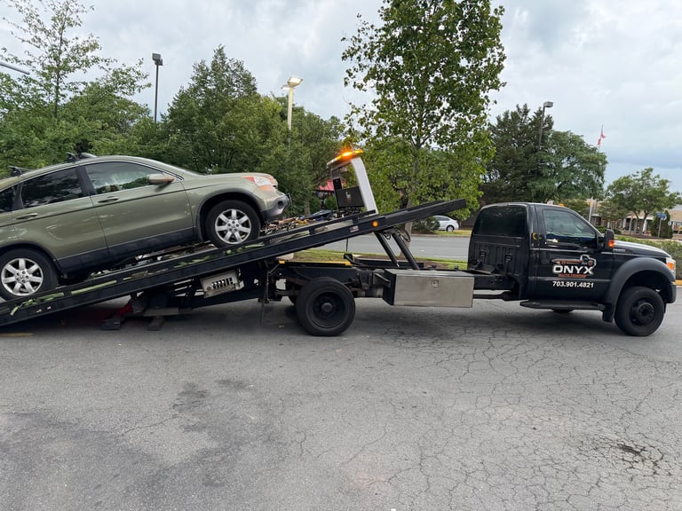 Black tow truck lifting a silver sedan onto its flatbed in a parking lot with trees in the background