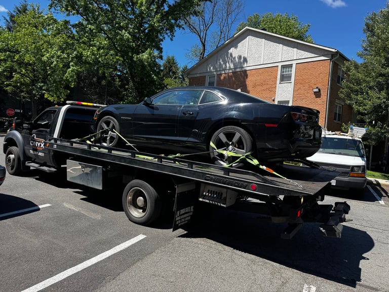 Black tow truck carrying a black sports car on its flatbed in front of a brick house