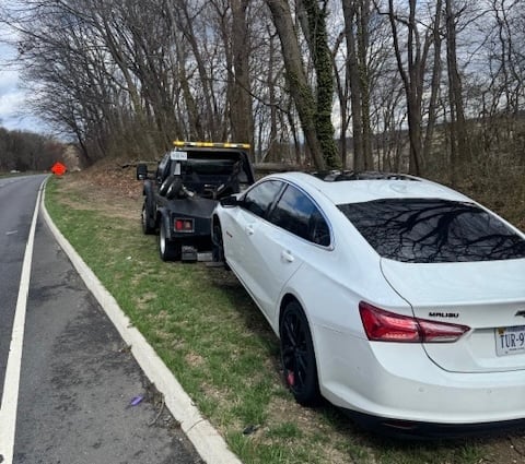 White Mitsubishi sedan being towed by a black flatbed truck on a rural road lined with bare trees