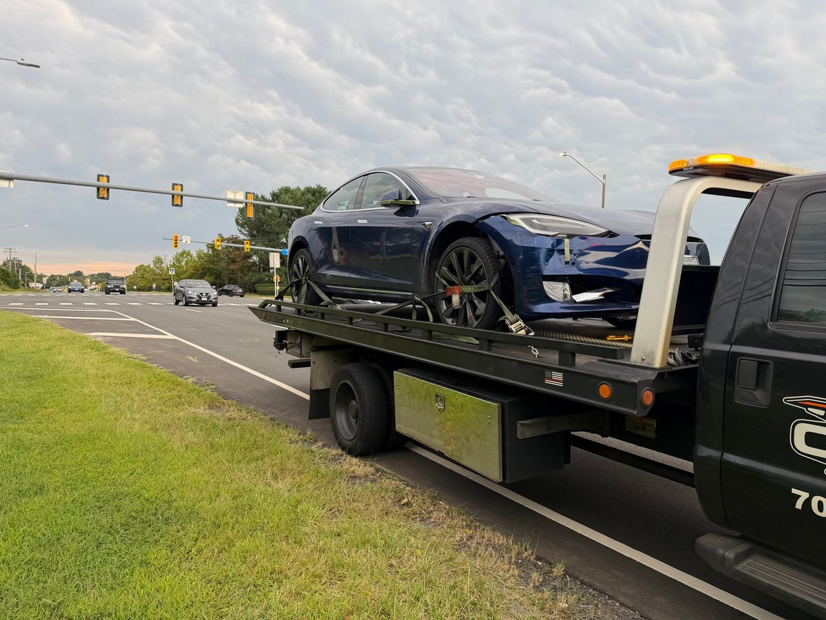 A blue Tesla Model 3 being towed on a flatbed truck along a highway on a cloudy day.