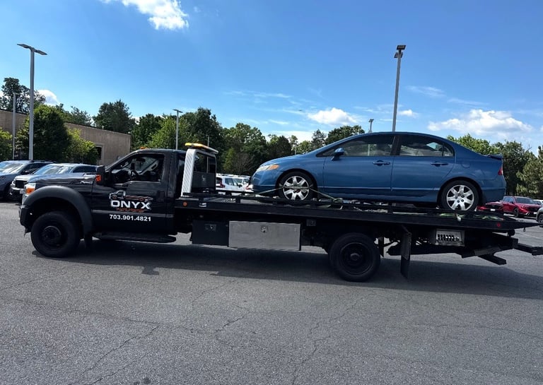 Black tow truck with open driver door transporting a blue sedan on its flatbed trailer in a parking lot