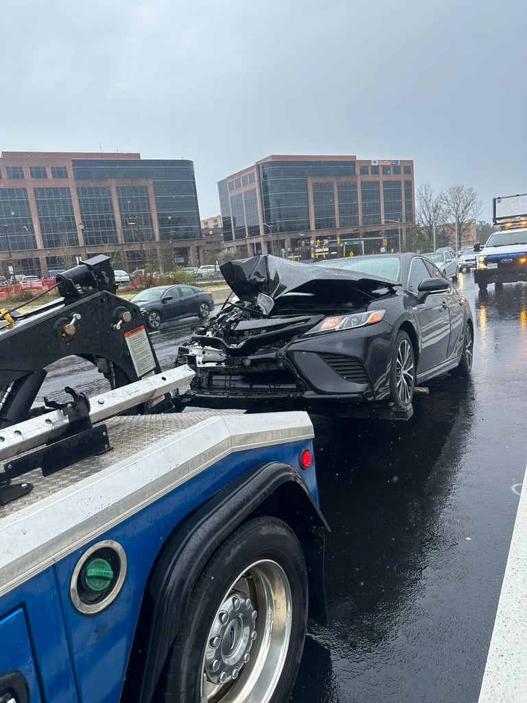 Damaged gray sedan on blue and white tow truck on wet highway with buildings in background